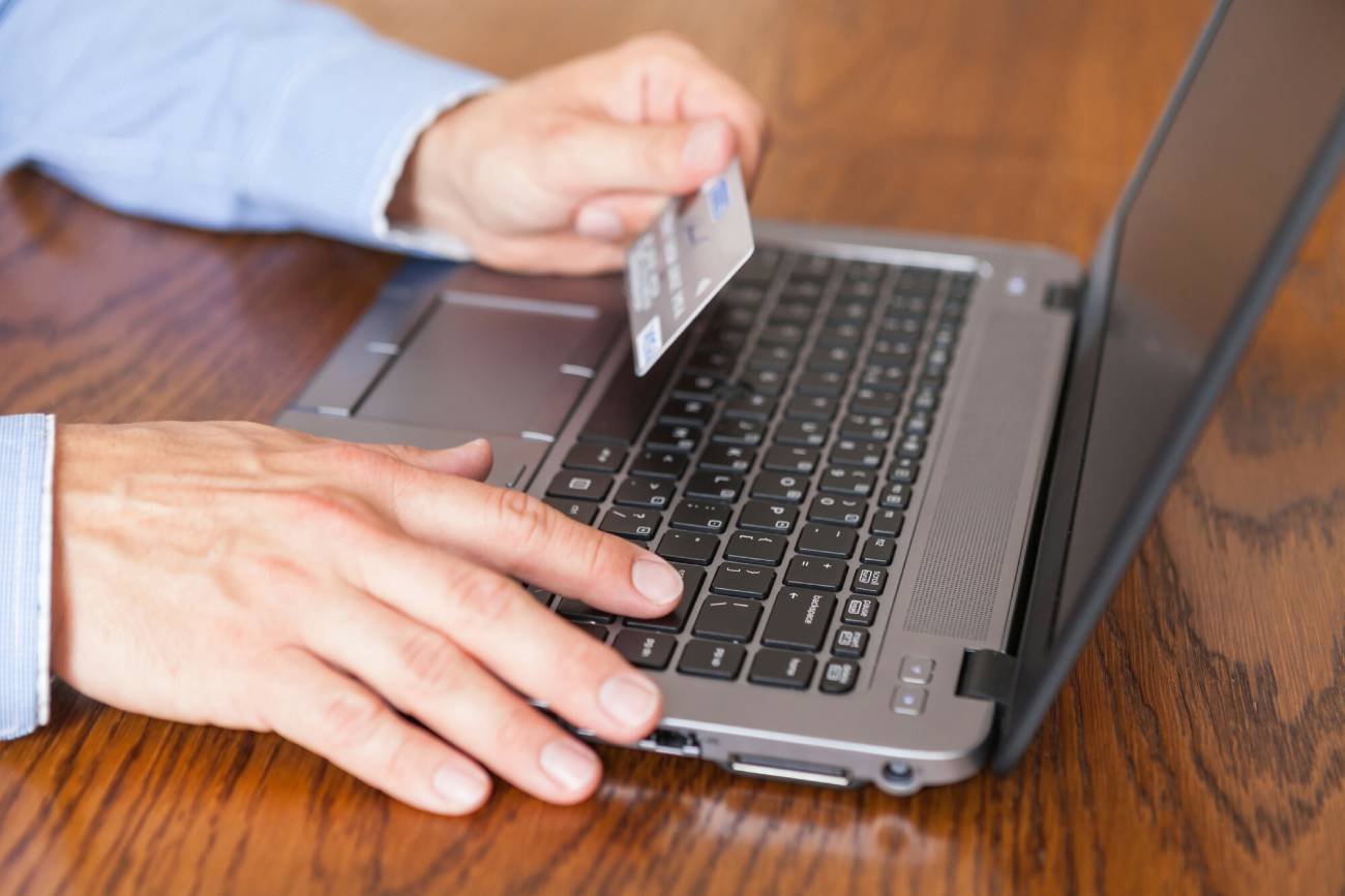 Mężczyzna trzyma kartę płatniczą przed ekranem laptopu.