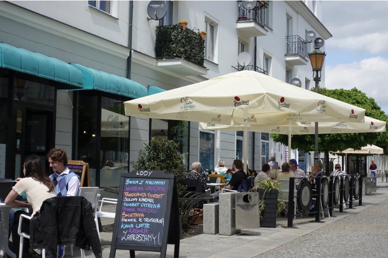 Ogródek gastronomiczny na ulicy w polskim mieście. Ludzie siedzą przy stolikach pod dużymi parasolami, na pierwszym planie znajduje się również tablica z wypisanym menu.