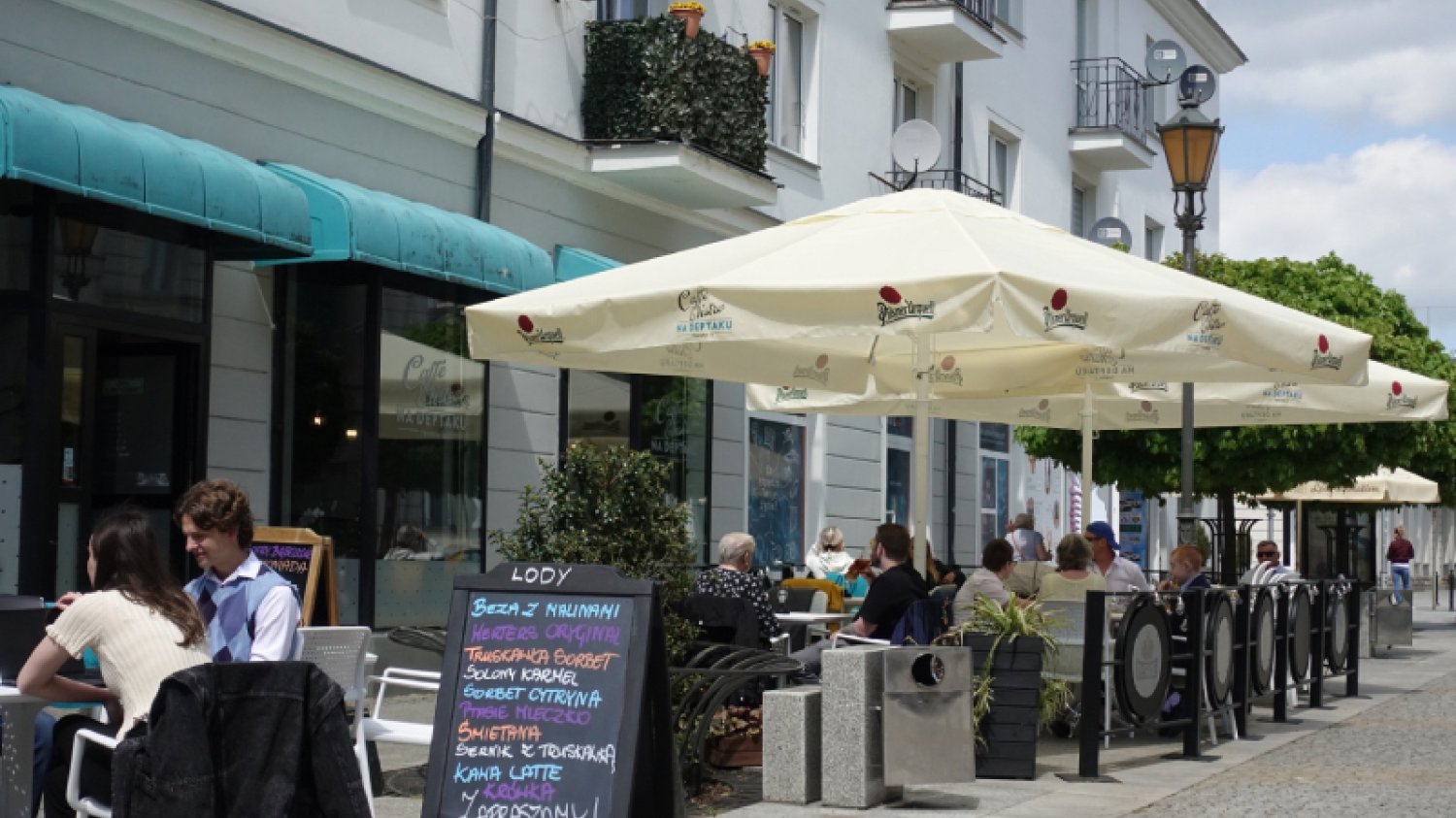 Ogródek gastronomiczny na ulicy w polskim mieście. Ludzie siedzą przy stolikach pod dużymi parasolami, na pierwszym planie znajduje się również tablica z wypisanym menu.