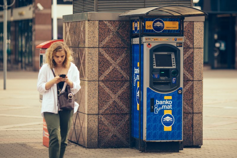 Przy ścianie stoi niebieski bankomat. Obok niego przechodzi młoda kobieta spoglądająca w telefon.