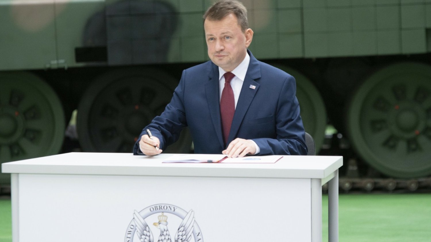 Mariusz Błaszczak przy biurku na tle czołgu