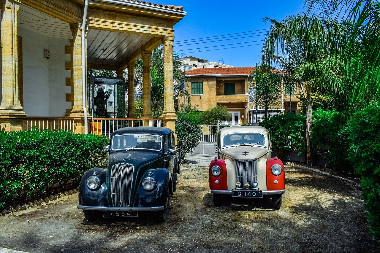 Cypryjska słoneczna ulica z palmami i piaskowymi budynkami, na której stoją dwa samochody old-timery: niebieski i czerwony. W tle widać kawałek błękitnego nieba.