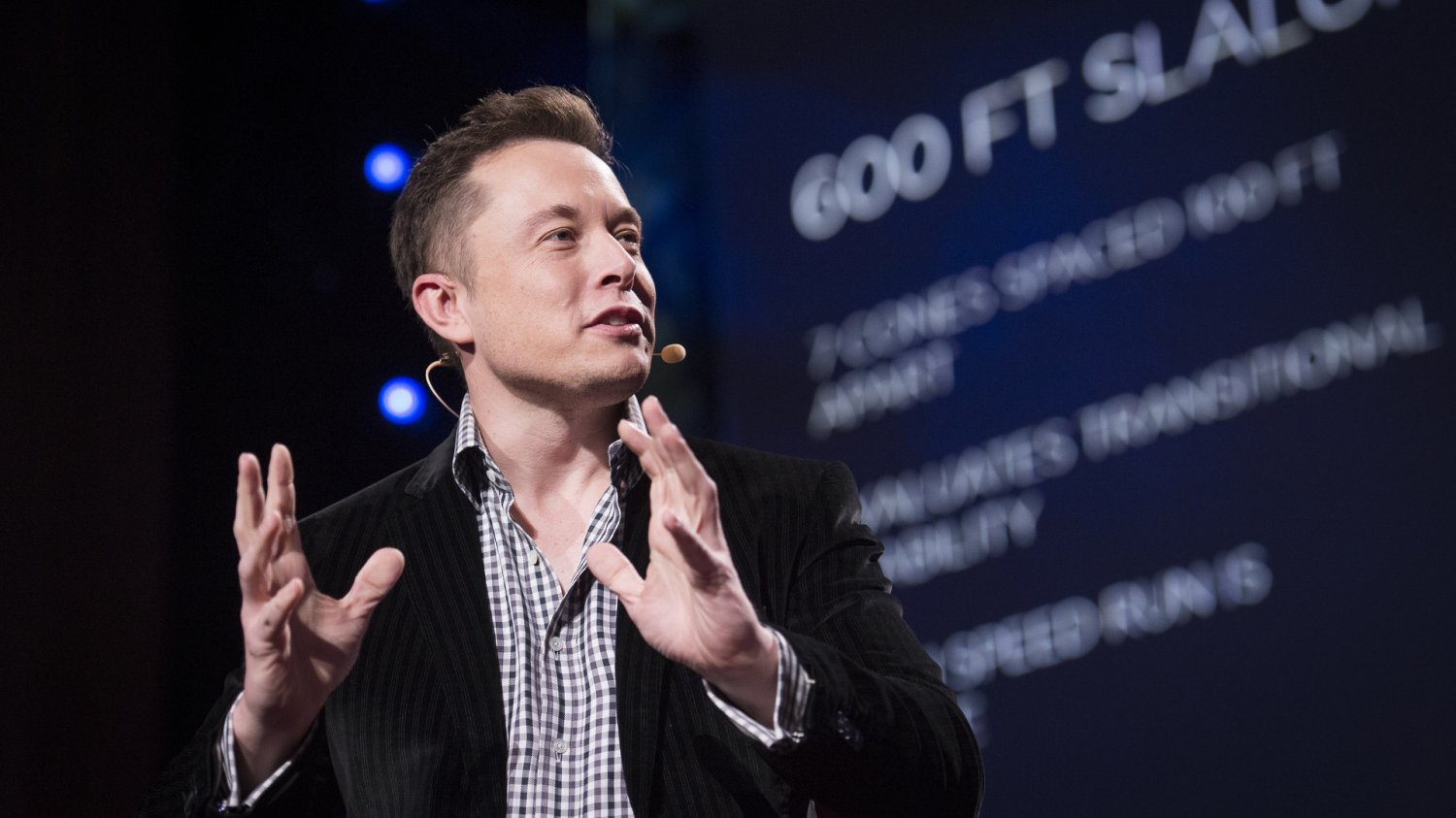 Elon Musk siedzący w czarnej marynarce i gestykulujący dłońmi