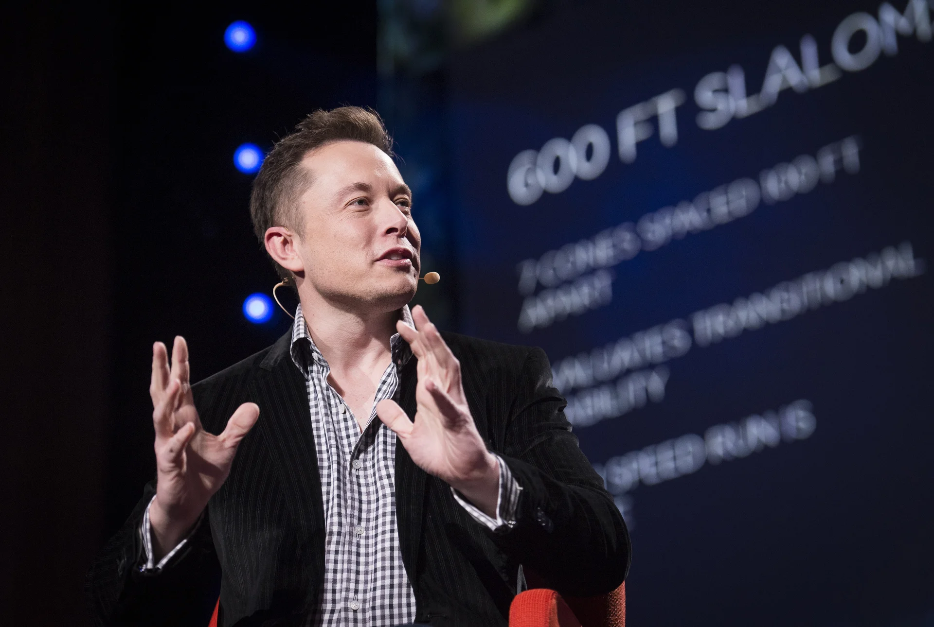 Elon Musk siedzący w czarnej marynarce i gestykulujący dłońmi