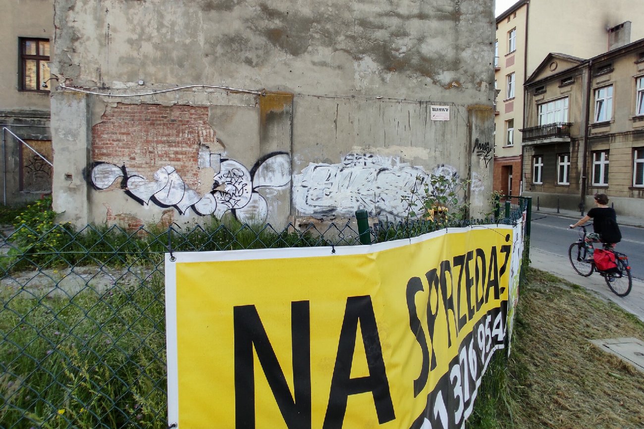 ogłoszenie na sprzedaż wisi na płocie kamiennicy obok którego przejeżdża kobieta na rowerze ubrana  na czarno