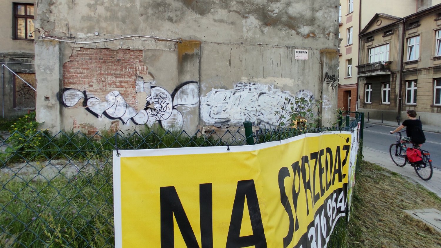 ogłoszenie na sprzedaż wisi na płocie kamiennicy obok którego przejeżdża kobieta na rowerze ubrana  na czarno