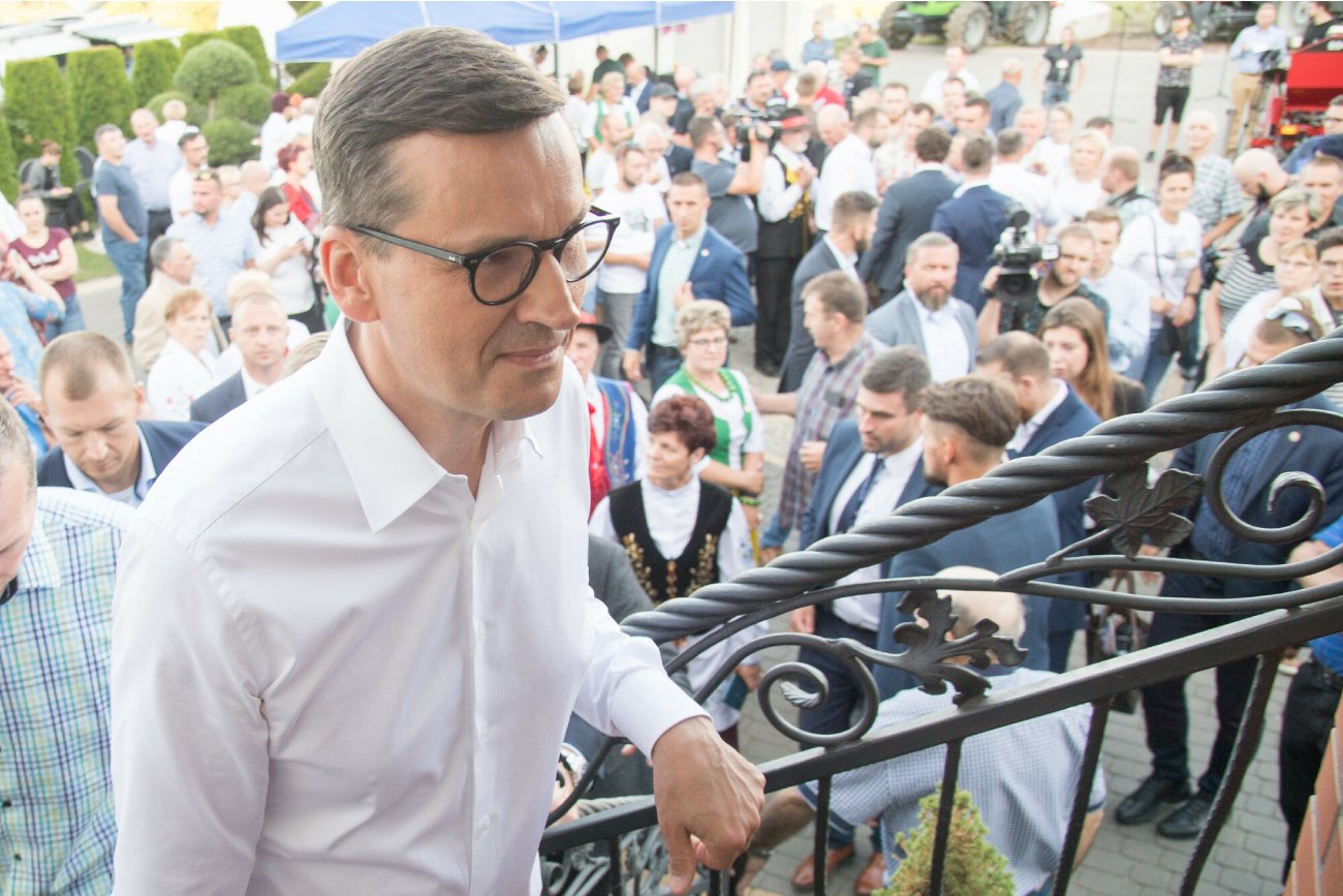 Mateusz Morawiecki w białej koszuli wchodzi o schodach na tle tłumu i trzyma się mosiężnej balustrady