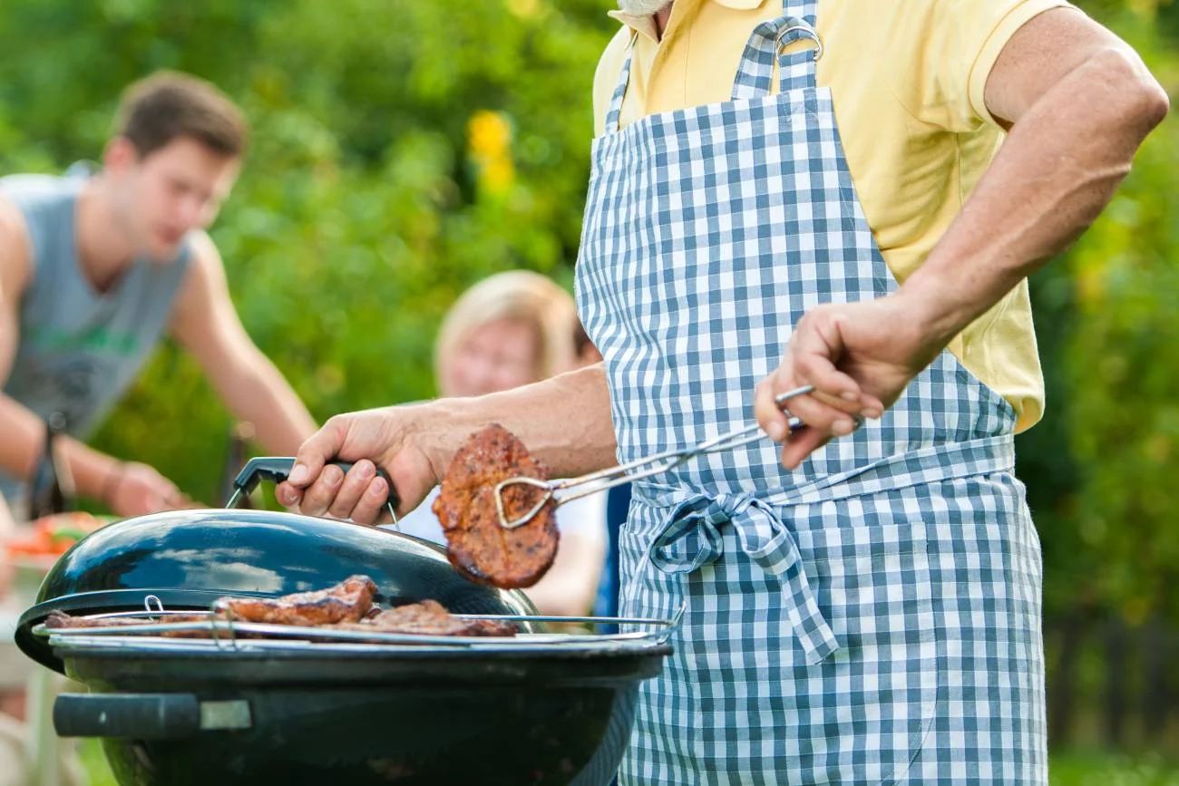 Starszy mężczyzna przy grillu przygotowuje karkówkę. W tle rodzina w ogrodzie.