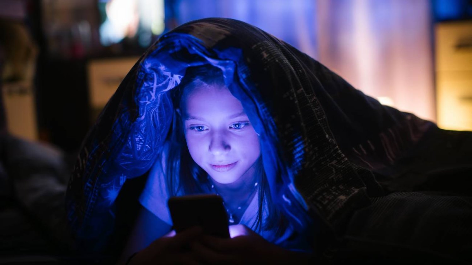 Młoda dziewczyna przegląda aplikacje na telefonie siedząc pod kołdrą.