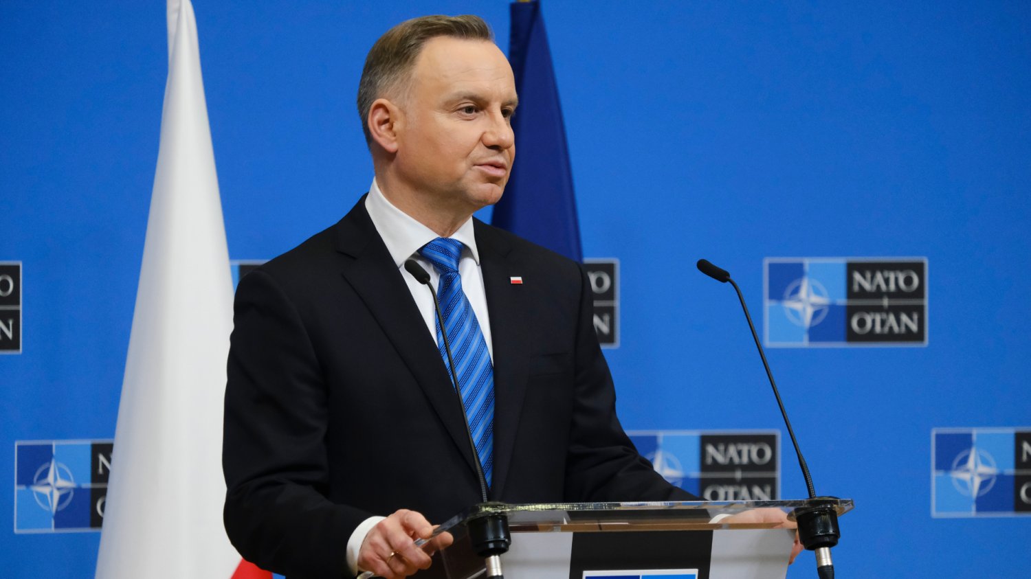 Andrzej Duda znalazł pracę w fundacji w USA. Sława Heritage Foundation ją wyprzedza Andrzej Duda przemawia. W tle niebieska ścianka z logiem NATO i dwie flagi. Flaga Polski i flaga NATO.