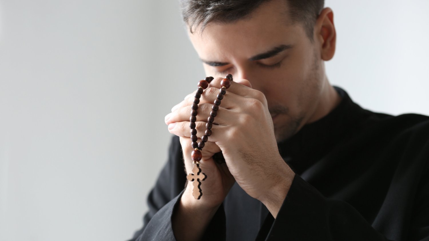 młody ksiądz z różańcem modlący się na jasnym tle