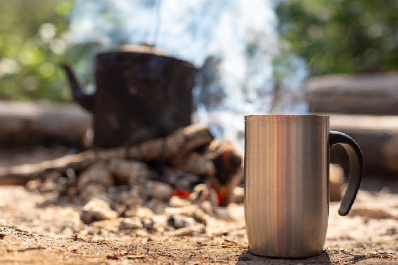 srebrny kubek termiczny z czarną rączką stojący na piaskowej ziemi przy ognisku