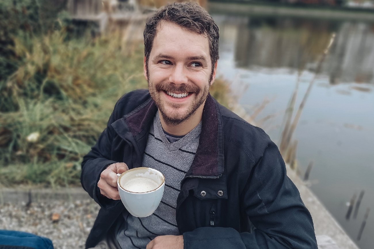 młody mężczyzna z brodą siedzi nad jeziorem z filiżanką kawy w ręku
