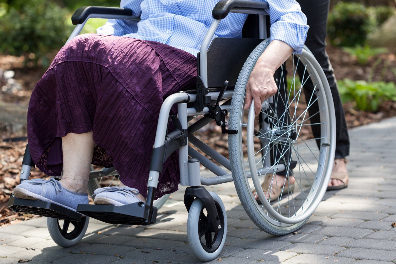 Starsza kobieta na wózku inwalidzkim na spacerze w parku w słoneczny dzień. Za nią jest młodsza kobieta, która prowadzi wózek.