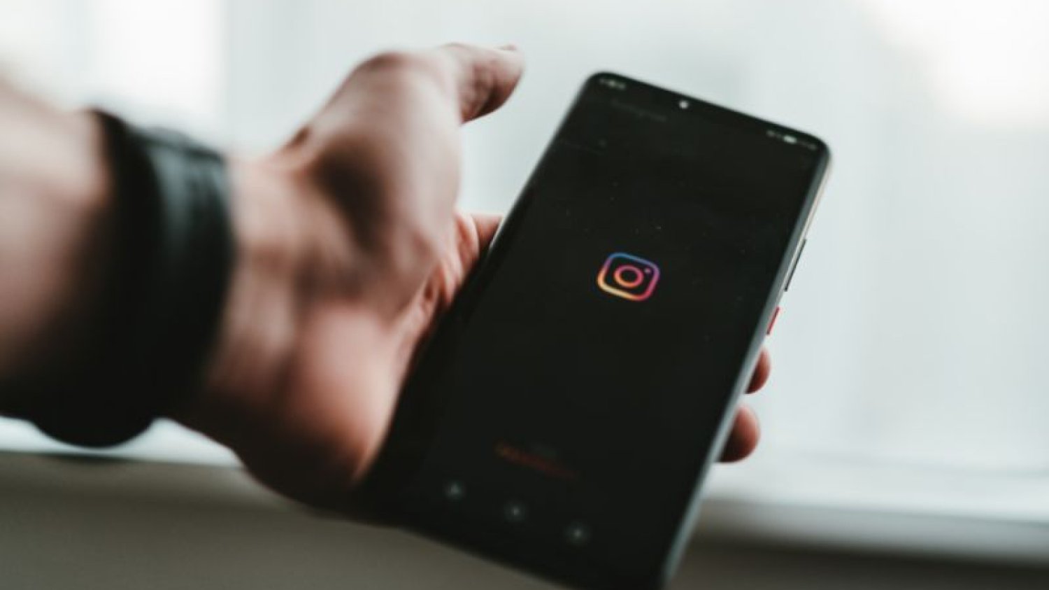 Smartfon, na którym wyświetla się logo Instagrama, trzymany przez męską dłoń z zegarkiem.