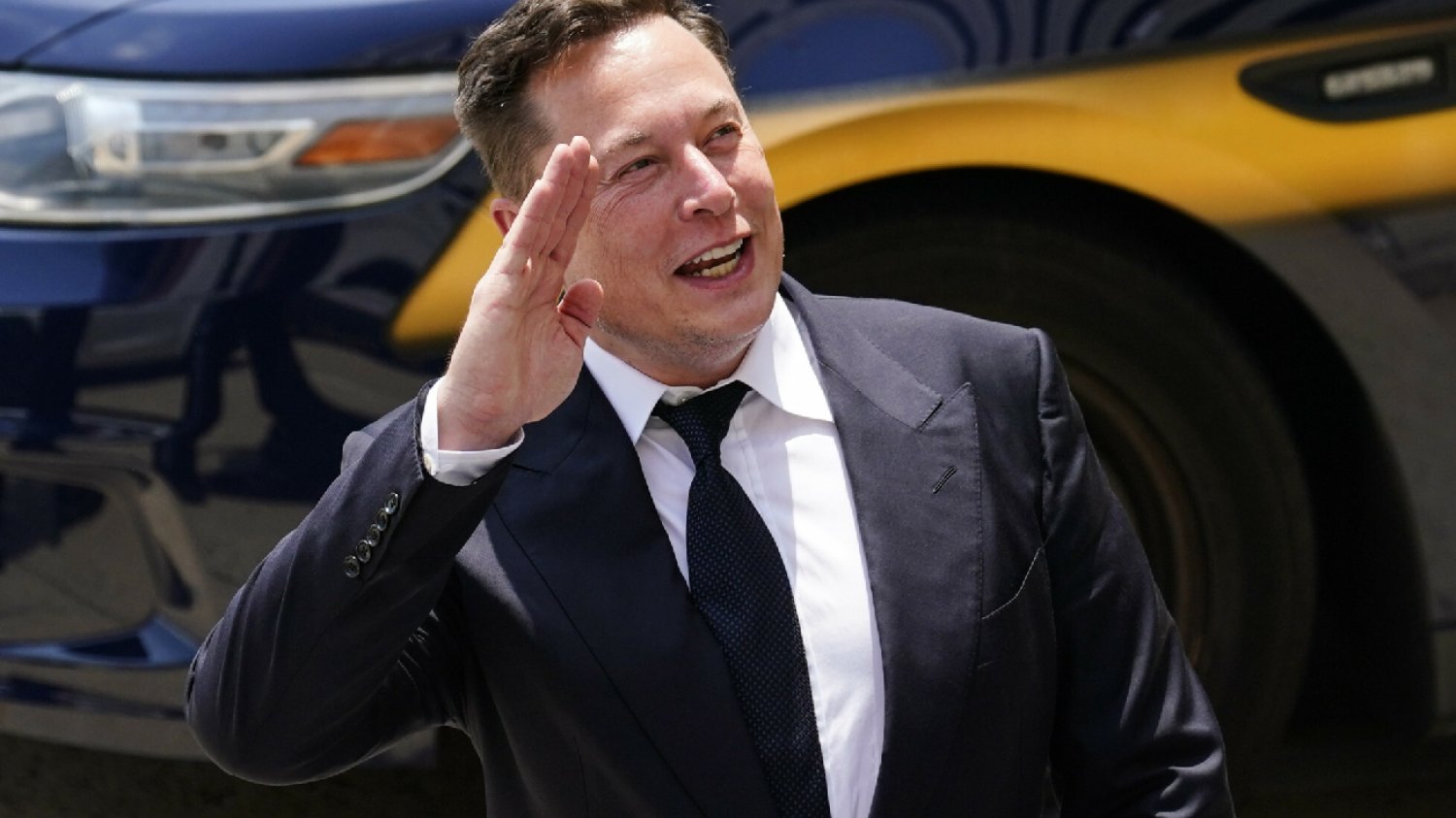 Elon Musk macha ręką stojąc na tle niebieskiego auta