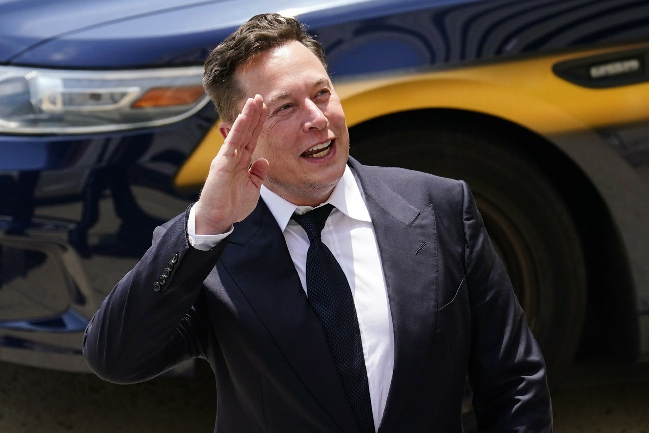 Elon Musk macha ręką stojąc na tle niebieskiego auta