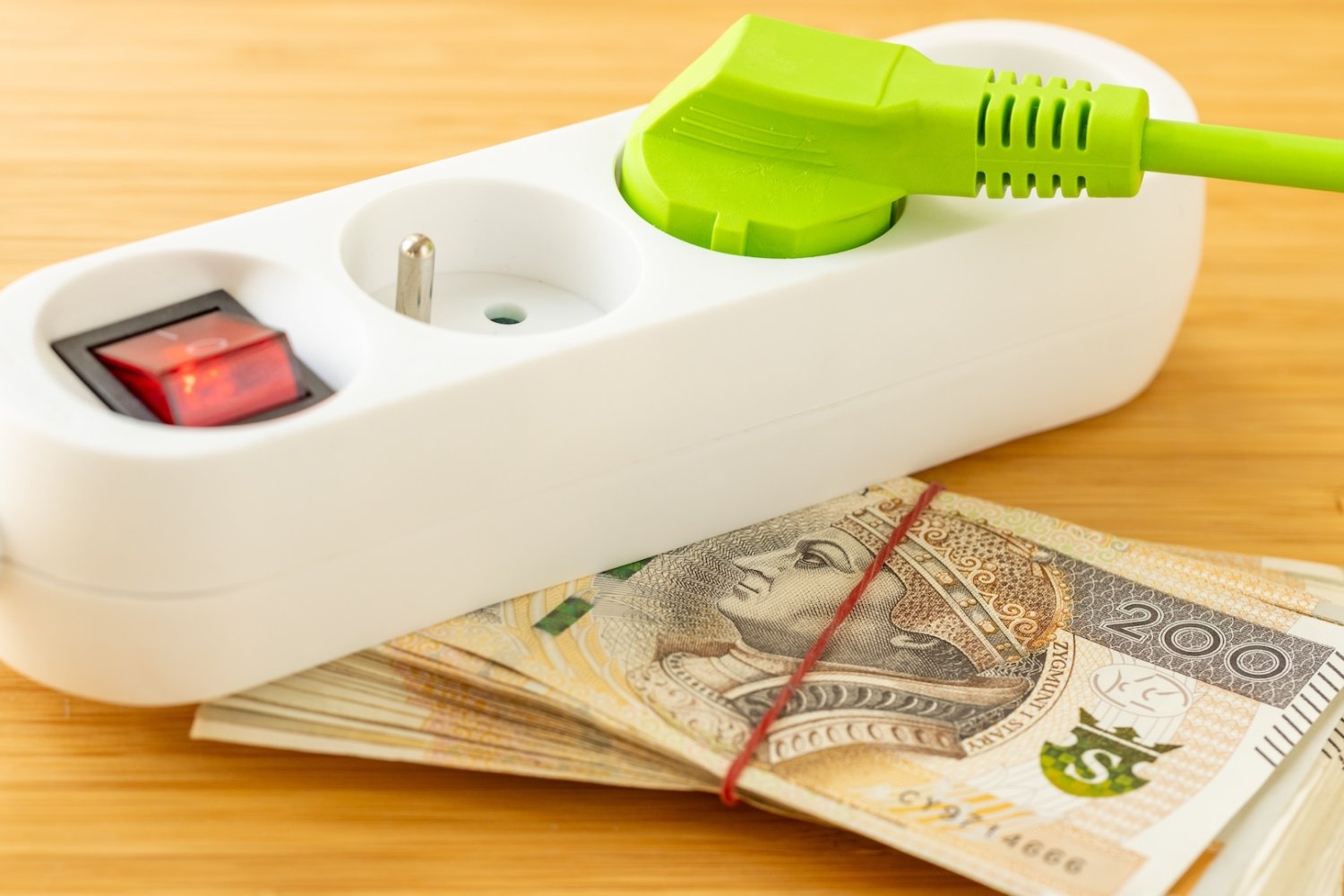 Rachunki za prąd w Polsce spadną? Niemcy mają plan na tanią energię. Listwa z podłączoną wtyczką elektryczną, a pod nią stos banknotów 200 zł.