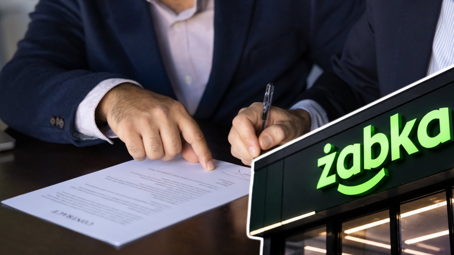 Dwóch mężczyzn podpisuje kontrakt. Sklep z logo Żabka w prawym dolnym rogu.